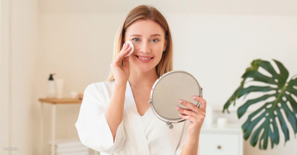 Woman in a bathrobe (MODEL) cleansing her face with a cotton round